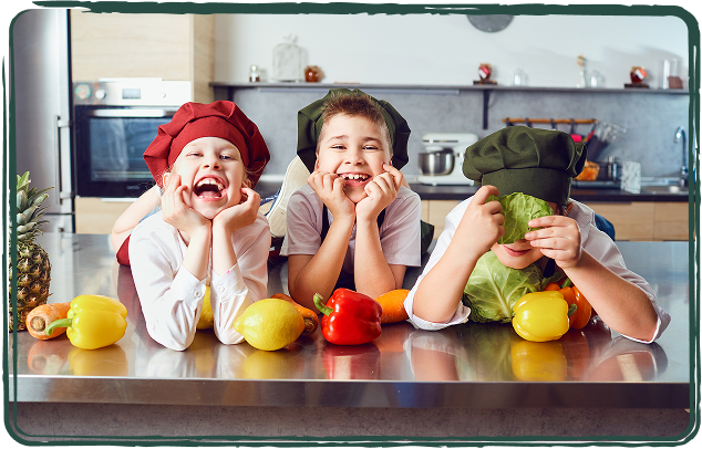 Drei Kinder mit Kochmützen stehen hinter einer Küchenarbeitsfläche mit Gemüse